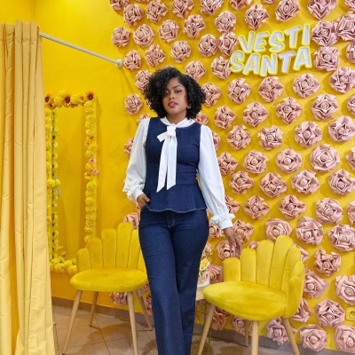 Mulher com roupa de ganga azul e camisa branca posando em frente a parede amarela com rosas cor de rosa e texto VESTI SANTA