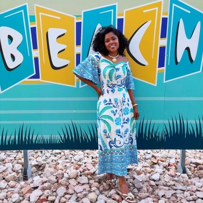 Mulher com vestido comprido floral azul e verde e sandálias douradas em frente a painel colorido com texto