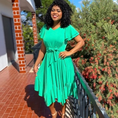 Vestido verde com mangas curtas e babados, cinto na cintura, usado por mulher numa varanda com ladrilhos vermelhos e plantas ao fundo