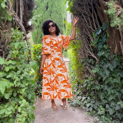 Mulher com vestido floral laranja e branco e óculos escuros num caminho com vegetação