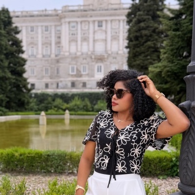 Mulher com top preto com flores brancas, calças brancas, óculos de sol castanhos, pulseira dourada, junto a um lampião em parque com palácio ao fundo