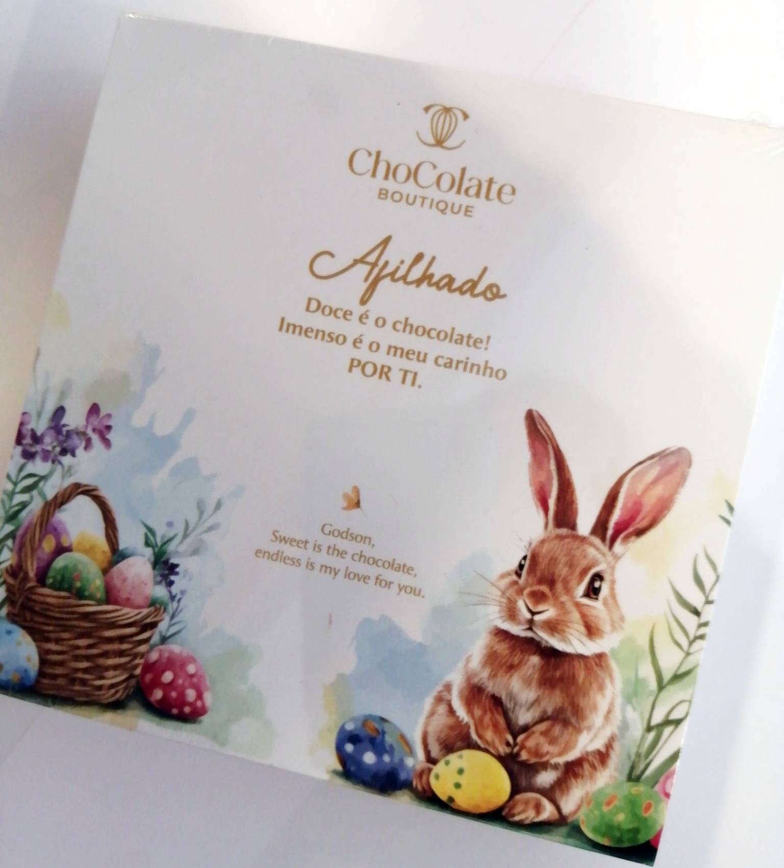 Maria Chocolate Caixa 9 Bombons Afilhado Chocolate Fabrico Artesanal Embalagem branca com texto dourado, coelho e ovos de Páscoa coloridos.
