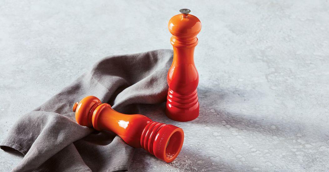 Le Creuset Set Mini Moinho Sal e Pimenta Utensílios Dois moedores de especiarias em madeira laranja sobre pano cinzento e superfície cinzenta.