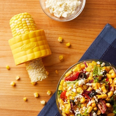 Espiga de milho com suporte amarelo ao lado de salada colorida numa tigela de vidro sobre mesa de madeira