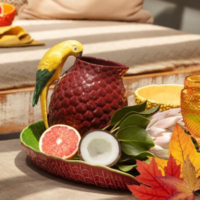 Jarro cerâmico vermelho com pássaro amarelo e verde numa bandeja com frutas e folhas