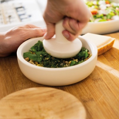 Almofariz cerâmico branco com ervas frescas a ser esmagadas, sobre superfície de madeira.