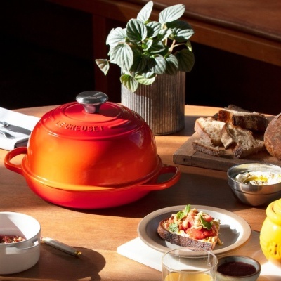 Panela vermelha Le Creuset com tampa metálica sobre mesa de madeira com comida e planta