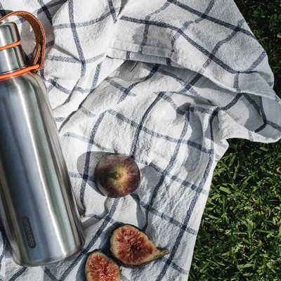 Garrafa térmica em inox com tampa prateada e pega castanha sobre pano branco com padrão azul, figos e relva