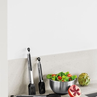 Utensílios de cozinha em inox e plástico preto com salada num tacho de inox e frutas sobre bancada de pedra