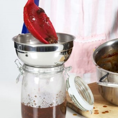 Frasco de vidro com molho a ser vertido por funil inox a partir de concha vermelha