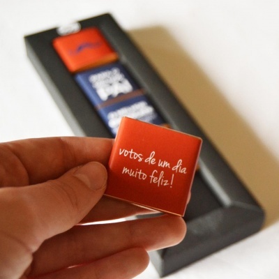 Chocolate quadrado com mensagem em embalagem vermelha sendo segurado, caixa preta com chocolates ao fundo