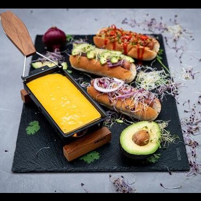 Três cachorros-quentes com diferentes toppings e queijo derretido em recipiente ao lado, abacate cortado ao meio e cebola roxa, tudo sobre uma tábua preta.