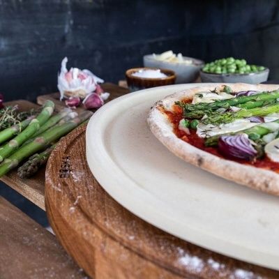 Pizza vegetariana com espargos e cebola roxa sobre pedra cerâmica branca em suporte de madeira.