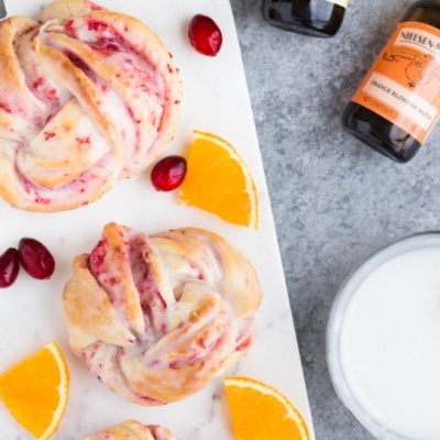 Pães doces com frutos vermelhos e laranja, jarra com bebida, garrafa com rótulo laranja em fundo cinzento