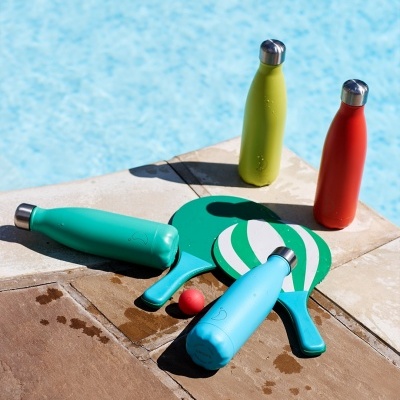 Garrafas isolantes coloridas junto a raquetes e bola à beira de piscina