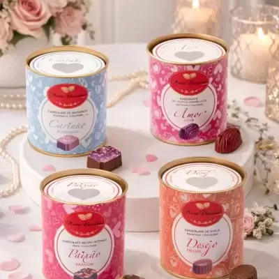 Quatro latas coloridas de chocolates Maria Chocolates sobre mesa branca com chocolates, flores e velas.