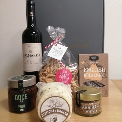 Produtos gourmet com vinho tinto, bolachas, chá, compota, queijo e patê sobre mesa de madeira clara.