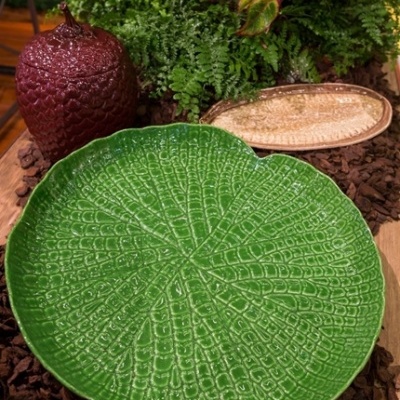 Prato decorativo verde em cerâmica com padrão de folhas e objetos decorativos ao fundo
