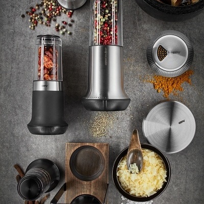 Moedores de especiarias em metal e vidro com especiarias coloridas e tigelas pequenas em fundo cinza.