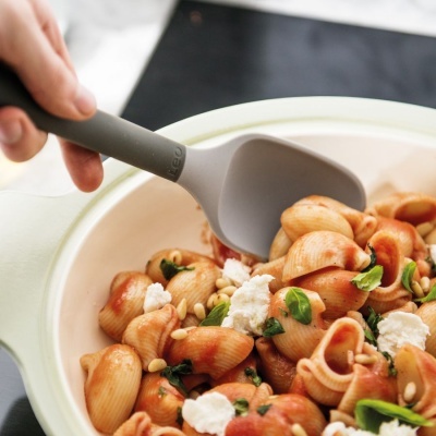 Tigela branca com massa concha em molho vermelho, queijo, folhas verdes e pinhões, e colher cinzenta.