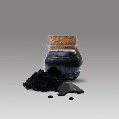 Frasco de vidro com tampa de cortiça com líquido preto e pó preto à sua frente sobre superfície cinzenta