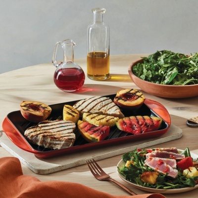 Tabuleiro com bifes e pêssegos grelhados, garrafas, salada e talheres numa mesa de madeira.