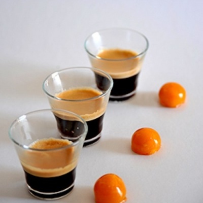 Três copos pequenos com café expresso e três frutos laranja em linha sobre fundo branco.