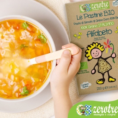 Pacote de massa Le Pastine BIO Alfabeto junto a tigela de sopa com massa em forma de letras e mão segurando colher