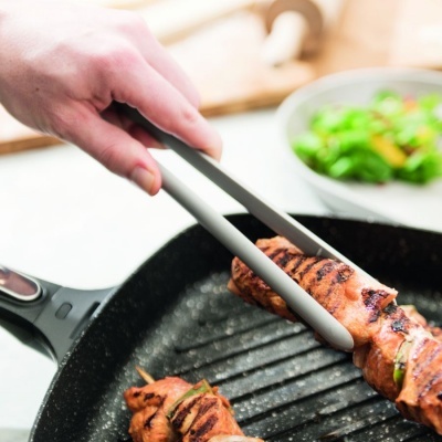 Prego de carne grelhada num tabuleiro de ferro fundido com pinças de cozinha e salada ao fundo