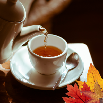 Chaleira a servir chá numa chávena branca com pires e colher, folhas de outono ao lado