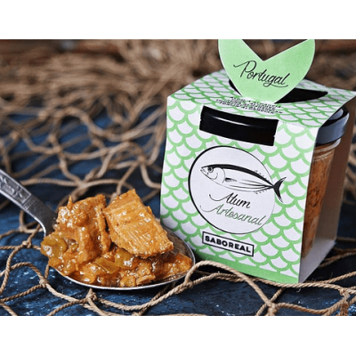 Frasco de vidro com atum artesanal Saboreal ao lado de uma colher com atum em pedaços sobre tecido azul e rede de pesca marrom