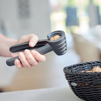 Quebra-nozes cinza escuro sendo usado para abrir noz junto a cesta de vime preta com mais nozes