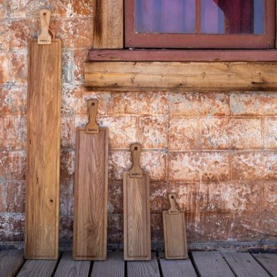 Quatro tábuas de madeira para cozinha, de vários tamanhos, encostadas numa parede de tijolos vermelhos junto a uma janela.