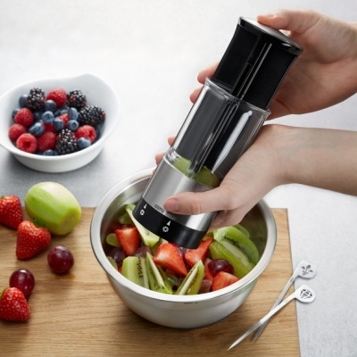 Moedor elétrico preto e cinza com frutas numa tigela metálica e frutas frescas ao lado