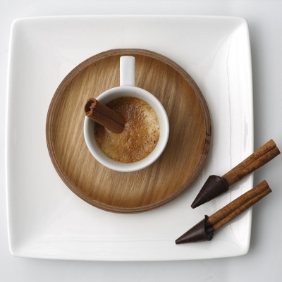 Chávena pequena com café e pau de canela numa base de madeira e prato branco, com pau de canela decorado com chocolate ao lado.