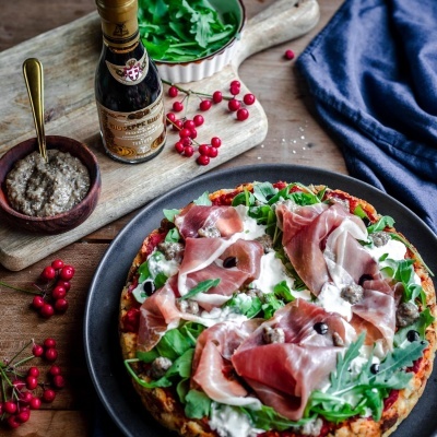 Pizza com presunto, rúcula e queijo em prato preto com azeite e decoração rústica