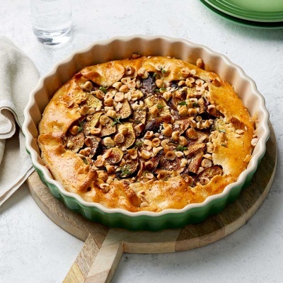 Tarte em forma cerâmica branca e verde com frutos secos e figos, sobre tábua de madeira, pano branco e garrafa de vidro