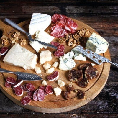 Tabuleiro redondo de madeira com queijos, enchidos, nozes e facas, sobre mesa rústica escura