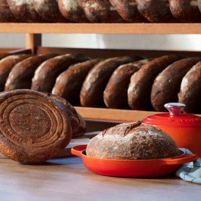 Pão redondo com crosta rija numa travessa vermelha e pães semelhantes em prateleiras de madeira ao fundo