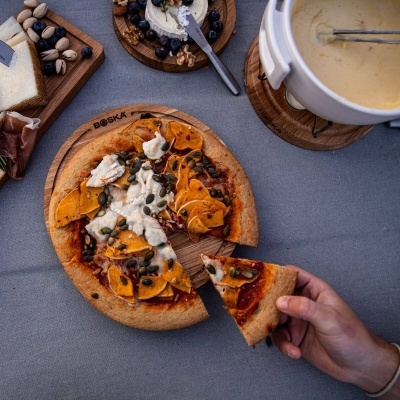 Pizza com queijo e sementes sobre tábua de madeira BOSKA com mão a pegar uma fatia