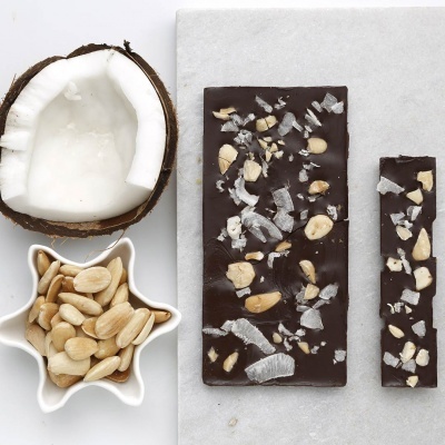 Barra de chocolate negro com amendoins e coco junto a coco partido e amendoins em recipiente