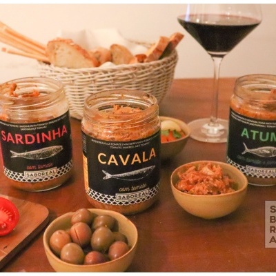 Três frascos de patê de peixe com rótulos pretos numa mesa com azeitonas, tomate, pão e copo de vinho