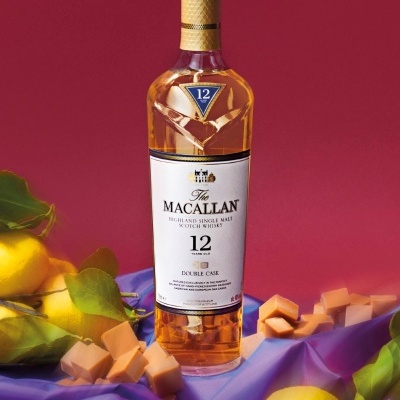 Garrafa de uísque The Macallan 12 anos com queijos e limões num pano lilás e fundo vermelho