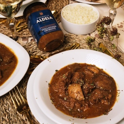 Pratos brancos com estufado de vitela, frasco de Conservas Aldeia, arroz, vinho branco e talheres dourados numa mesa decorada.