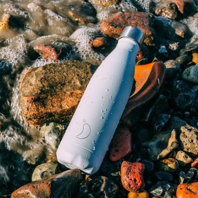 Garrafa térmica branca com gotas de água sobre pedras coloridas à beira de água