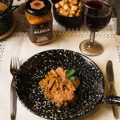 Frasco de molho entrecosto com lentilha, prato de comida com lentilhas, copo de vinho, tigela com croutons em mesa com toalha clara.