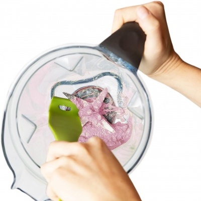 Liquidificador com líquido rosa e espátula verde, mãos seguram o copo e mexem.