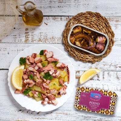 Salada de polvo com batata e limão servida num prato branco com lata de polvo, limão e azeite ao lado sobre mesa rústica.