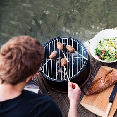 Grelhador portátil com espetos de carne, baguete, faca e salada na beira da água