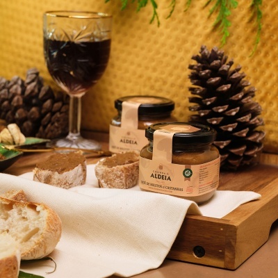 Frascos de vidro com produto cremoso rotulados 'ALDEIA', pão, copo de vinho tinto, pinhas e fundo amarelo.